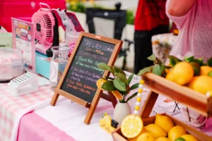 A market stall table displays lemons, a chalkboard menu, a pink fan, a payment terminal, and decorative items on a pink checkered tablecloth.
