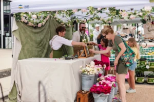 A woman and two girls buy flowers from a vendor at an outdoor market stall decorated with garlands and buckets of colorful flowers.