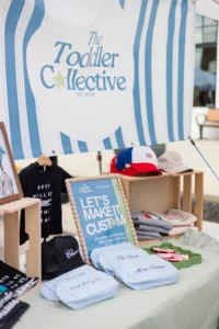A display table with toddler clothing, hats, and a sign reading "The Toddler Collective" and "LET'S MAKE IT CUSTOM" at an outdoor market booth.