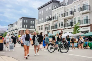 People walk and shop at an outdoor market on a city street lined with modern apartments; a cyclist rides by and tents are set up for vendors.