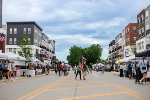 People walk along a closed street lined with vendor tents and modern buildings during an outdoor market event under a cloudy sky.