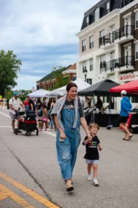 A woman and a young child walk hand-in-hand down a street lined with vendor tents at an outdoor market, with other people visible in the background.