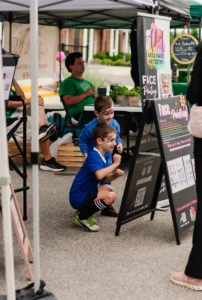 Two young boys with painted faces look at a face painting menu board at an outdoor event, surrounded by tents and seated adults.