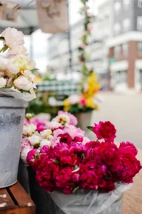 Buckets of pink and white peonies are displayed outside on a city sidewalk, with blurred buildings and street in the background.