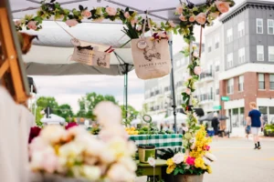 Flower-decorated market stall with Fables Farms tote bags, floral arrangements, and a green checkered tablecloth, set outdoors near apartment buildings.