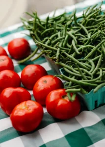 Several red tomatoes and a container of green beans sit on a green and white checkered tablecloth.