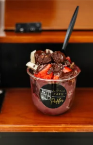 A cup of acai topped with sliced bananas, strawberries, brownies, cacao nibs, and chocolate sauce, with a black spoon inserted. The cup is labeled "Little Farm Peddler.