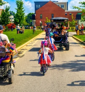 Children in festive outfits ride decorated bikes down a sunny street, followed by a golf cart, with adults and onlookers nearby during a community parade.