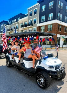 A woman and two children sit in a decorated golf cart with American flags, parked on a street in front of modern apartment buildings.