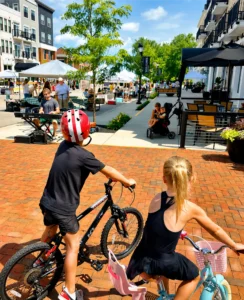 Two children on bikes face a lively outdoor market with tents, shoppers, and a musician performing on a brick-paved street lined with shops and restaurants on a sunny day.
