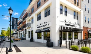 Street view of a modern building complex with shops, including an ice cream store, outdoor seating, and a "WELCOME" banner on a lamppost.