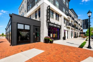Street view of a modern building with a sign reading “Sextons Pizza” and a large vertical “PIZZA” sign at the corner, with brick pavement and sidewalk in front.