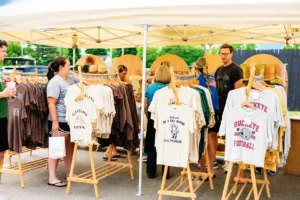 People browse t-shirts displayed on racks under a tent at an outdoor market. The shirts feature local and sports-related designs.