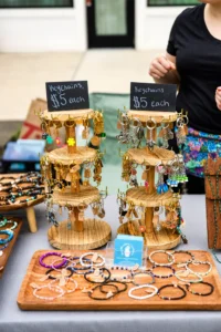 Wooden displays with assorted keychains priced at $5 each, alongside trays of beaded bracelets on a table at an outdoor market. A person stands behind the table.