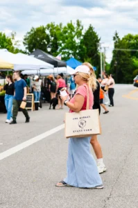 Woman holding a drink and market tote stands at an outdoor farmers market, with other people and vendor tents in the background.