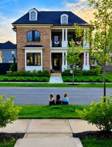 Three children sit on the grass across the street from a large brick house with white trim and patriotic decorations, during sunset.