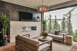Modern patio with a TV, fireplace, two sofas, a round table, potted plants, brick floor, and an open view of greenery and a house outside.