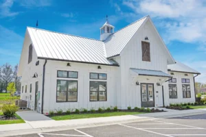 White modern barn-style building with a metal roof, large windows, and small garden beds, situated next to a paved parking lot under a partly cloudy sky.