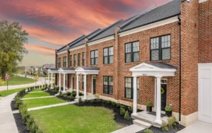 Row of modern red-brick townhomes with white columns and manicured lawns under a dramatic sunset sky.