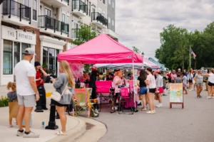 People gather at outdoor vendor tents on a street near modern apartments; a pink tent is prominent. Some visitors stand, chat, or walk while others browse items for sale.