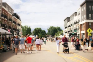 People stroll along a blocked-off street lined with market stalls and modern apartment buildings under a partly cloudy sky.