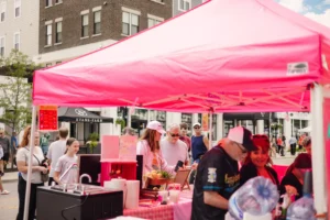 People gather under a bright pink tent at an outdoor market, with various food items and supplies on a checkered table. Buildings and other attendees are visible in the background.