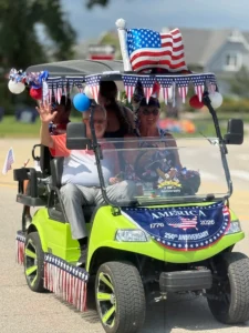 Four people ride a decorated green golf cart in a parade, with U.S. flags, red, white, and blue streamers, and a banner marking America’s 250th anniversary.