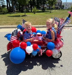 Two children sit in a red wagon decorated with American flags, balloons, and patriotic colors, outdoors on a sunny day.