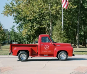 A vintage red pickup truck with "Evans Farm" logo is parked near a flagpole with an American flag. Trees and a clear sky are in the background.