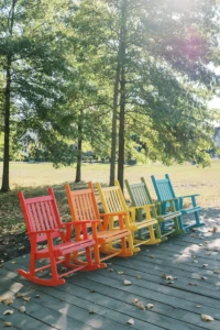 Six colorful rocking chairs, arranged in a rainbow pattern, sit on a wooden deck outdoors with green trees and sunlight in the background.