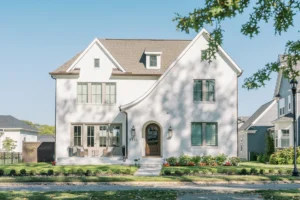 Two-story white brick house with arched wooden front door, large windows, front porch, and landscaped yard on a sunny day.