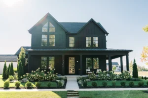 A modern two-story black house with large windows, a front porch, and landscaped yard featuring bushes, flowers, and a walkway leading to the front door.