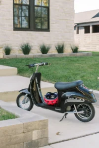 A black Razor electric scooter with a red helmet rests on the seat, parked on a concrete path beside a grassy yard and a stone house.