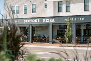 Street view of Sextons Pizza patio with several people seated at outdoor tables under a covered area; modern building exterior visible in the background.