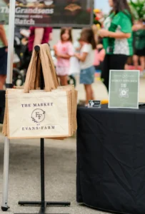 Reusable tote bags labeled “The Market at Evans Farm” hang on a stand beside a price sign at an outdoor market, with people and children in the background.