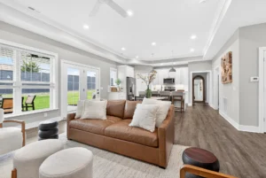 Modern living room with a brown leather sofa, neutral decor, open floor plan, large windows, and a view of the kitchen and dining area in the background.