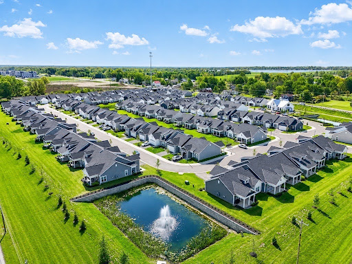 Aerial view of a suburban neighborhood with rows of houses, green lawns, a small pond with a fountain, and open fields in the background under a blue sky—an inviting glimpse for those looking to explore homeownership in Lewis Center.