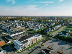 Aerial view of a suburban neighborhood with houses, roads, green lawns, parking lots, and commercial buildings under a clear blue sky.