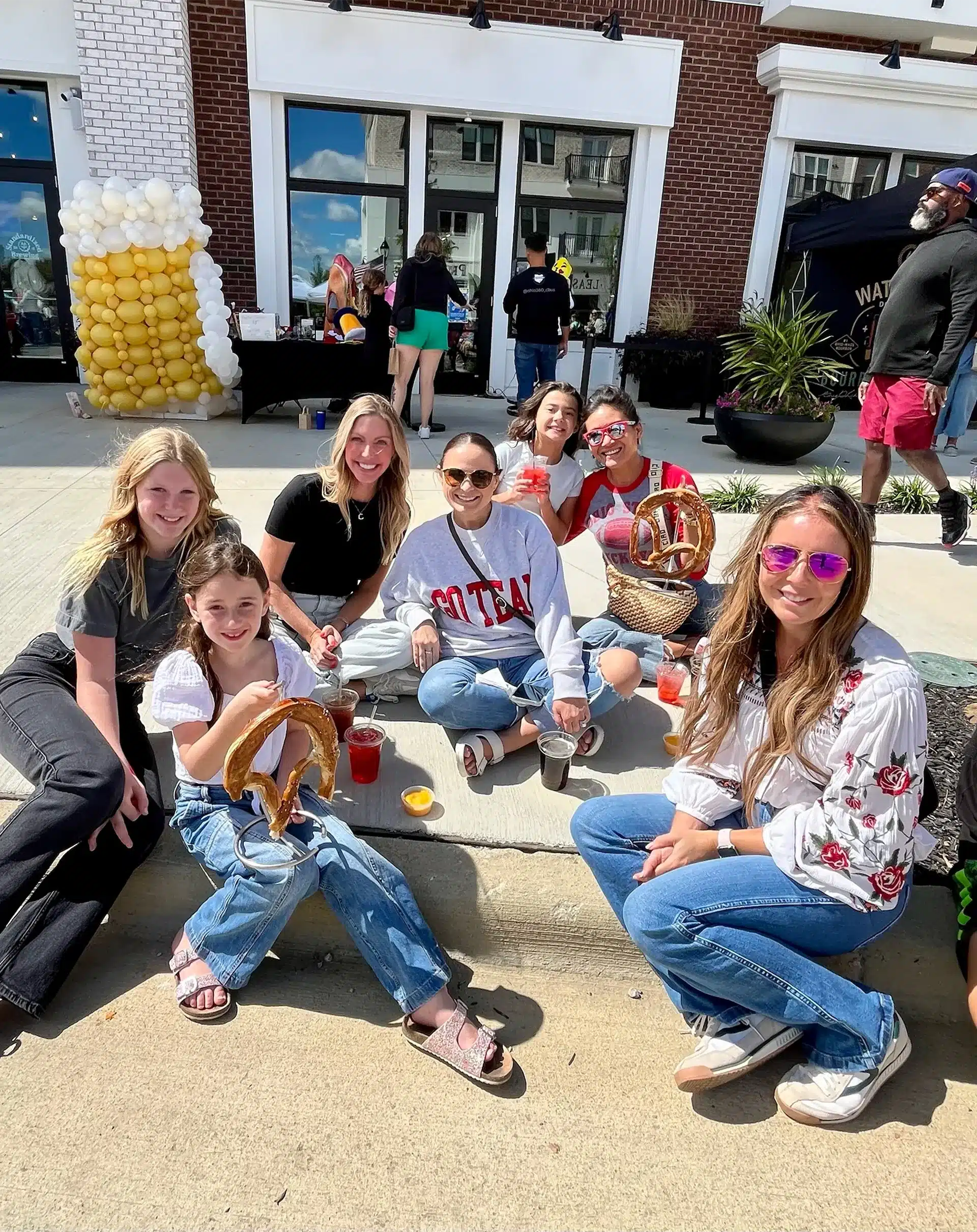 A group of six women sit on a sidewalk enjoying snacks and drinks on a sunny day, with event booths and decorations visible in the background.