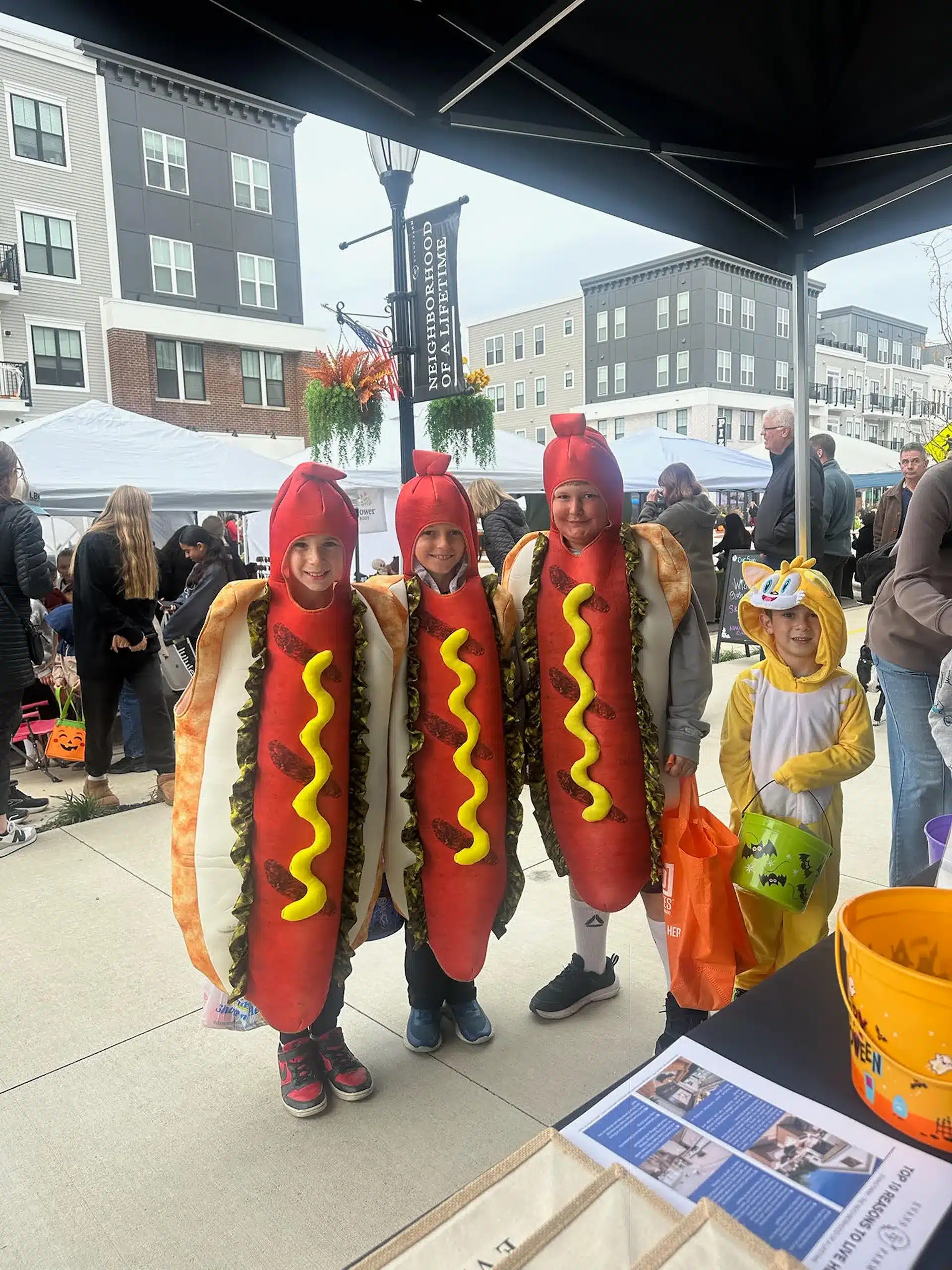 Four children in costume pose at an outdoor event; three are dressed as hot dogs, and one is dressed as a chicken. Apartment buildings and vendor tents are visible in the background.