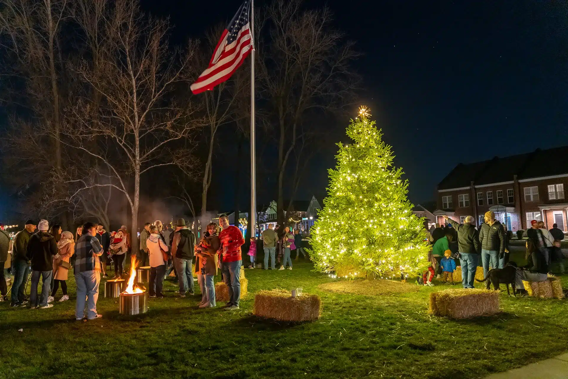 A group of people gather by a lit Christmas tree and a fire pit at night, with an American flag raised nearby and houses in the background.