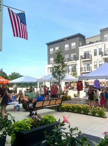 People visit outdoor market stalls with tents near modern apartments on a sunny day. An American flag is displayed above, and flowers line the sidewalk in the foreground, creating an inviting atmosphere to explore homeownership in Lewis Center.