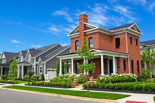 A row of suburban houses with manicured lawns, including a prominent red brick two-story home with a front porch under a blue sky—an inviting scene that invites you to explore homeownership in Lewis Center.