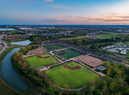 Aerial view of a sports complex with multiple baseball and softball fields, surrounded by trees, a pond, nearby roads, and residential areas at sunset—an inviting scene that perfectly captures the vibrant community where you can explore homeownership in Lewis Center.
