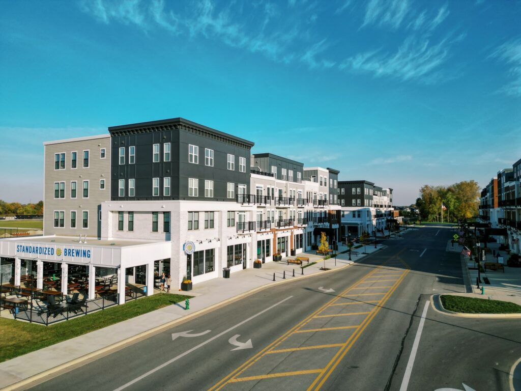 Modern mixed-use buildings with shops and apartments line a quiet street on a clear day; an outdoor seating area for a brewery is visible on the left.
