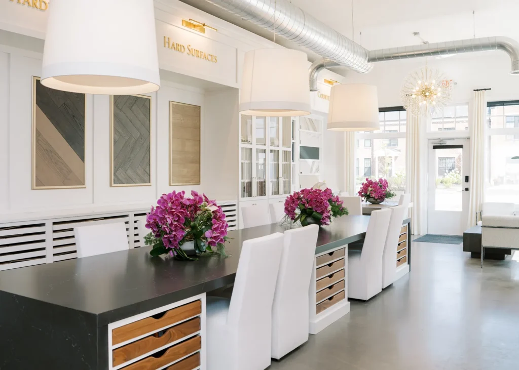 Modern showroom with long black tables, white chairs, large pendant lights, and purple flower arrangements. Wall samples and cabinetry are visible in the background. Discover Evans Farm Location - Ideal Living in Lewis Center, OH.
